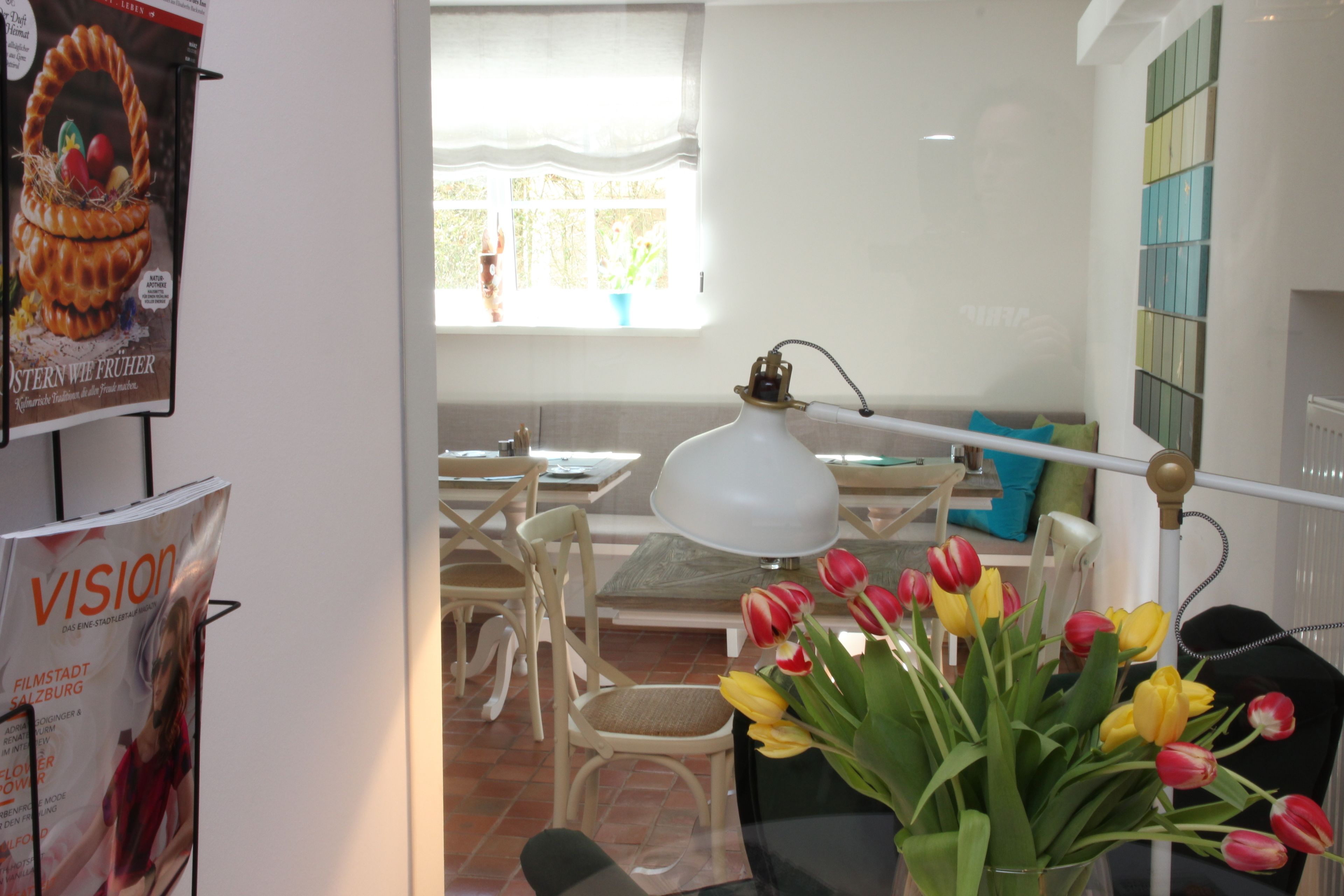Un comedor con una mesa, sillas y una lámpara. Hay una ventana con cortinas, una pared con un panel colorido y un cartel en la pared.