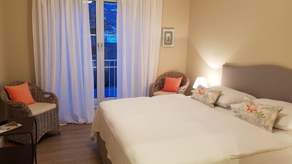 Habitación de hotel con una cama grande, dos sillones de mimbre, una mesita y una ventana con cortinas.