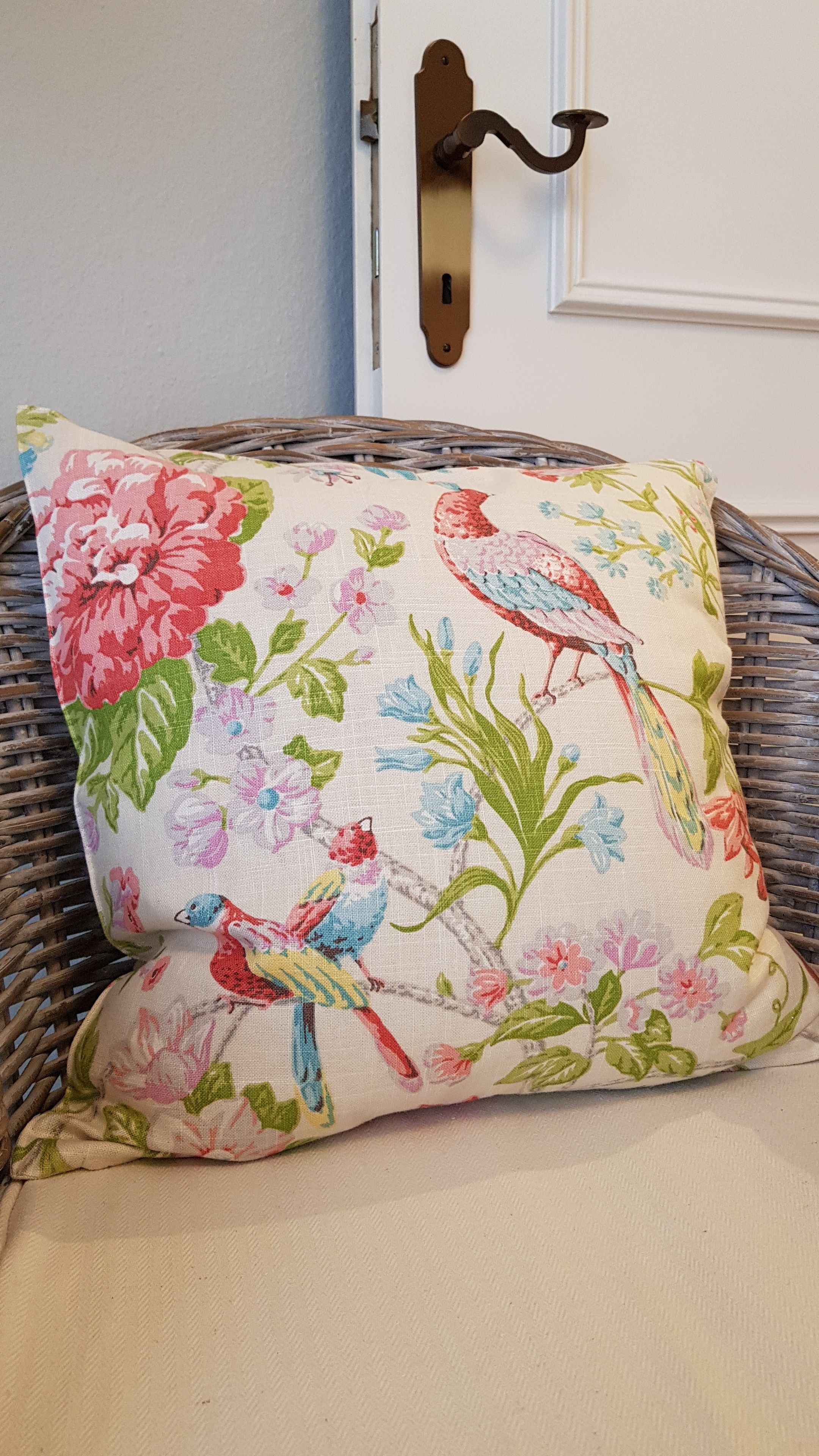 A cushion with a floral and bird pattern, placed on a wicker chair.