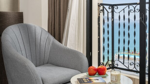 Una habitación con un sillón gris, una mesita redonda con un libro y dos manzanas, y vista al mar desde un balcón con barandilla negra.