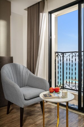 A room with a grey armchair, a small round table with a book and two apples, and a view of the sea through a balcony with a black railing. - Sochi Hotel (Nha Trang)