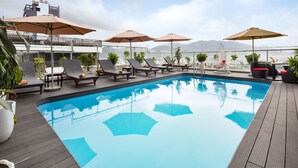 Una piscina en la azotea con sillas de descanso y sombrillas, rodeada de plantas en macetas y con vistas a un paisaje urbano.