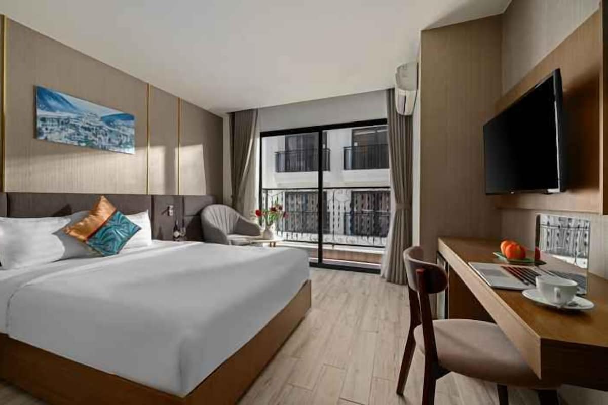 Habitación de hotel con cama, escritorio, silla, televisor y balcón con vistas.