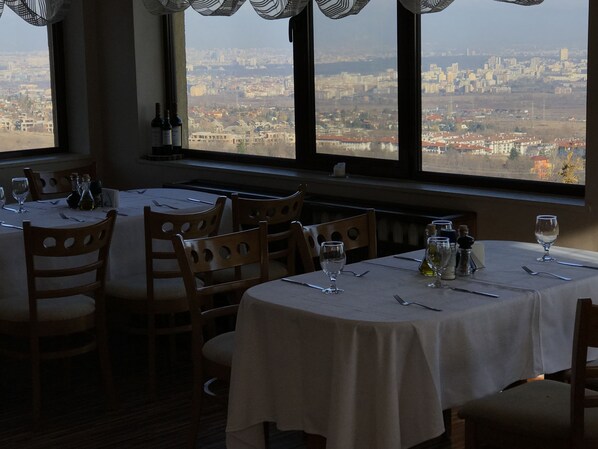 A restaurant with tables set for dining, a view of a cityscape through large windows, and bottles of wine on the windowsill. - Chairite (Sofia)