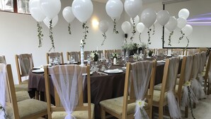 Une salle de banquet avec des tables rondes, des chaises recouvertes d’un tissu léger et transparent, des ballons blancs et des décorations florales.