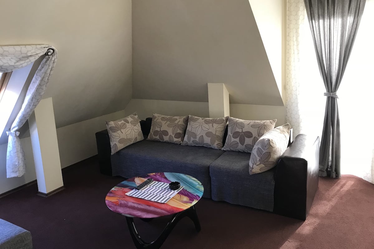 A cozy living room with a grey sofa, floral cushions, a colorful table, and a window with curtains.