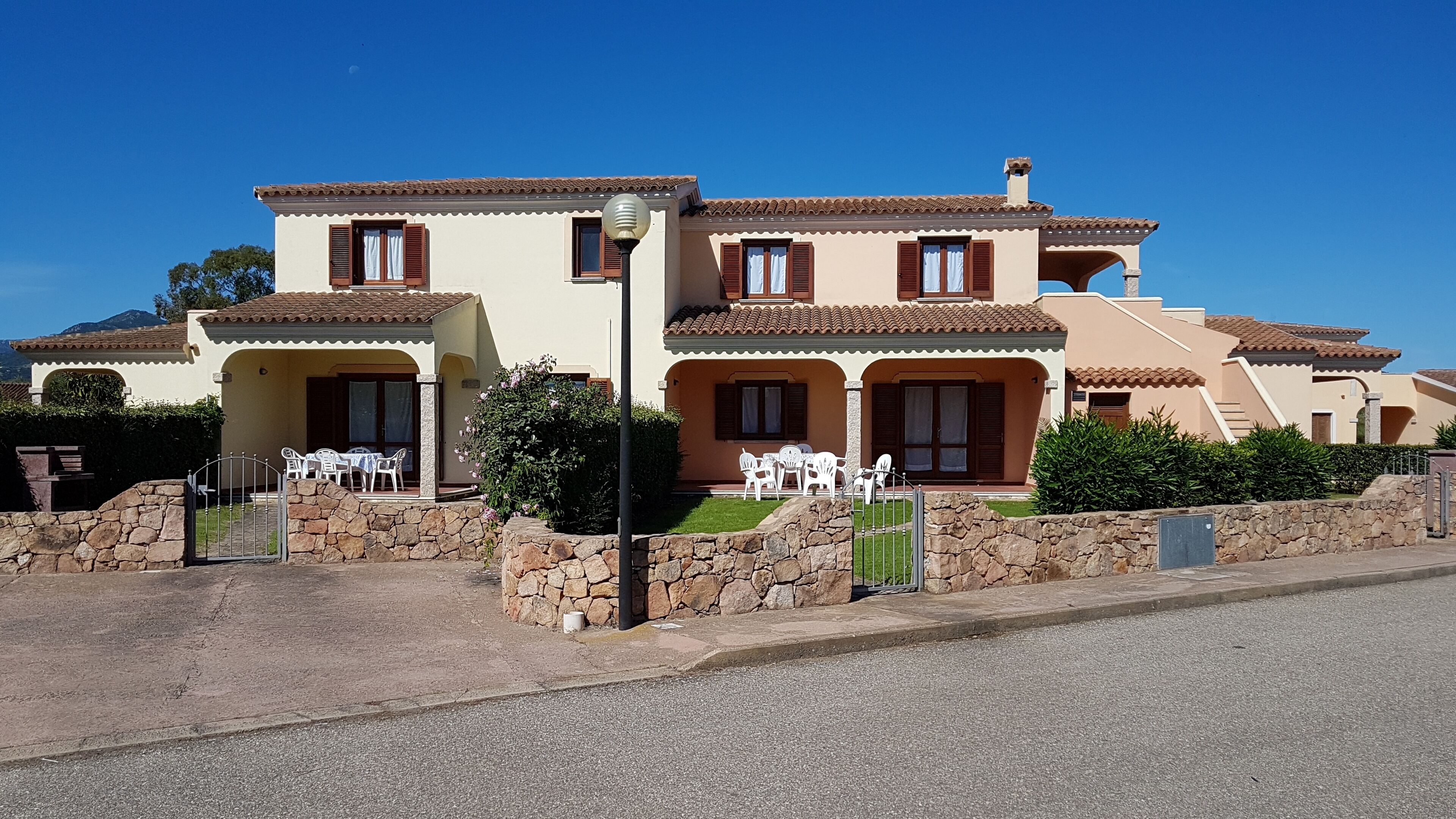 A two-story house with a stone wall, a patio with white chairs, and a clear blue sky.