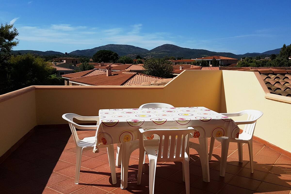 A balcony with a table and chairs, overlooking a residential area with houses and mountains.