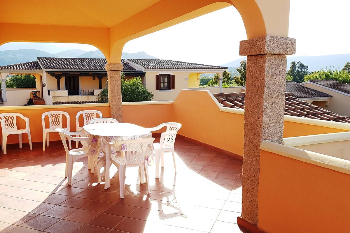 A covered outdoor seating area with a table and chairs, a stone pillar, and a view of buildings and mountains in the background.