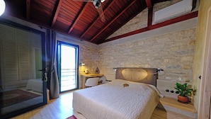 Ein Schlafzimmer mit einer Steinwand, Holzbalkendecke, einem Bett mit weißer Bettdecke, einem kleinen Tisch mit Lampe und einer Topfpflanze.