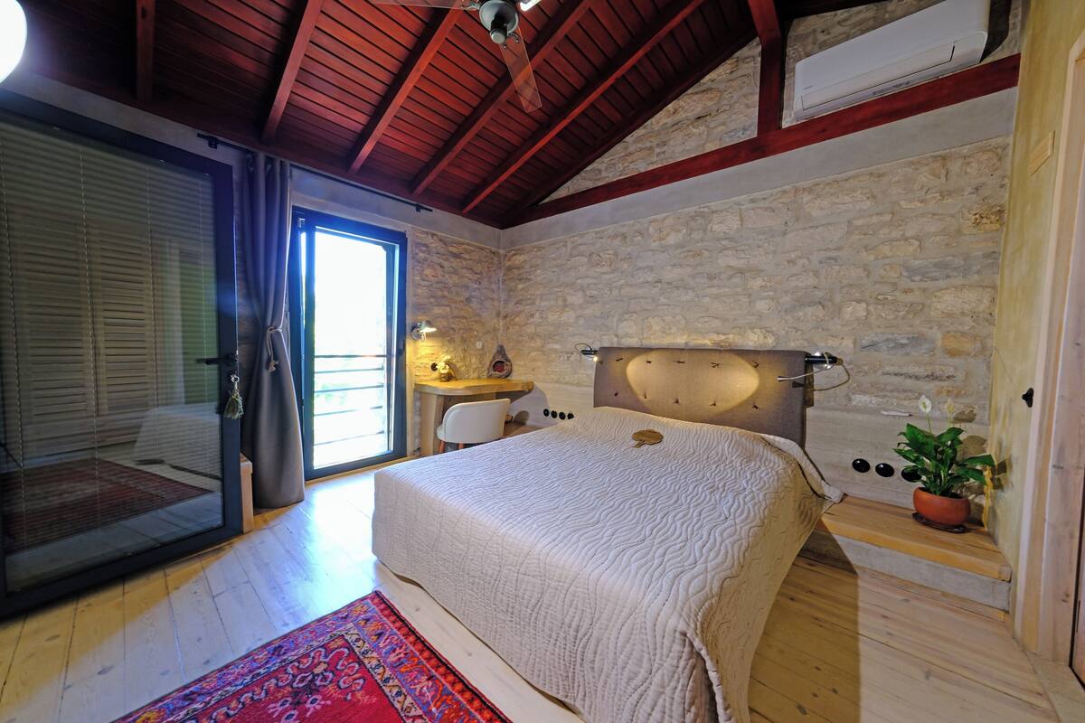 A bedroom with a stone wall, wooden ceiling beams, a bed with a white quilt, a small table with a lamp, and a potted plant.