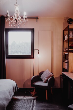 A modern bedroom with a large window, a chandelier, a chair, and a bookshelf. - At Home (Gesves)