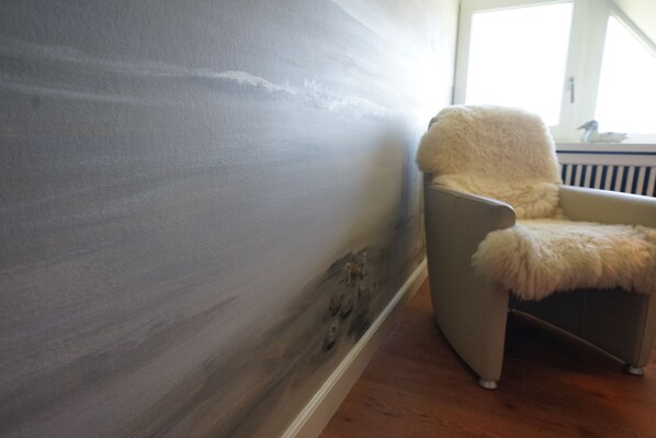 A room with a large window, a chair with a fluffy cushion, and a wall with visible damage. - Ferienwohnung Feskerdam 28 OG (Sylt)
