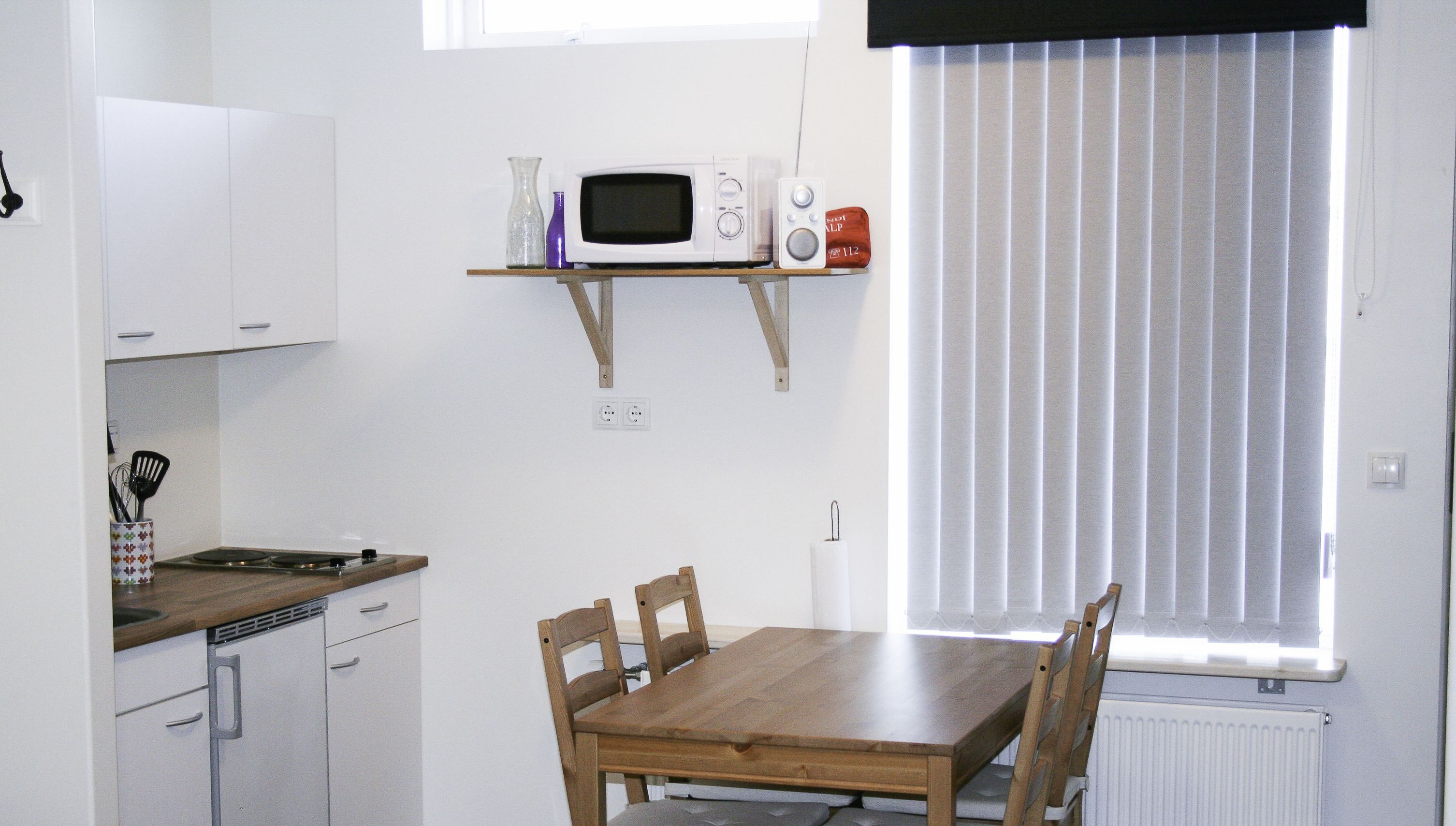 Una cocina compacta con microondas, un estante con adornos, una mesa de comedor con sillas y una ventana con persianas.