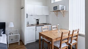 A compact kitchen with white cabinets, a wooden dining table, and chairs. There is a small side table with a lamp and a shelf above the sink holding a microwave and speakers. - South Central Apartments (Selfoss)
