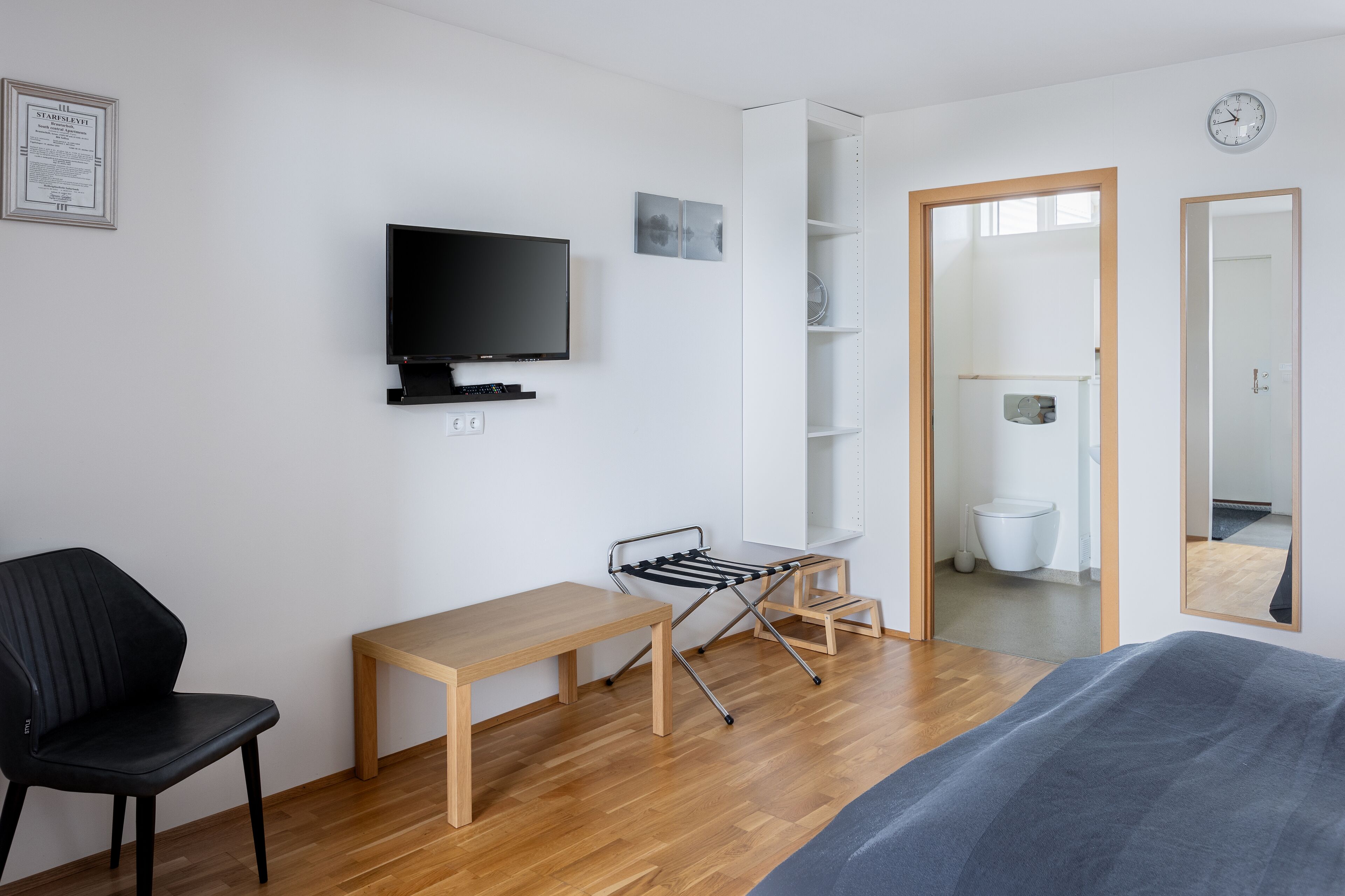 Quarto moderno e bem iluminado, com piso de madeira, uma cadeira preta, uma escrivaninha de madeira, uma televisão na parede, um varal e um espelho grande.