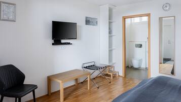 Quarto moderno e bem iluminado, com piso de madeira, uma cadeira preta, uma escrivaninha de madeira, uma televisão na parede, um varal e um espelho grande.