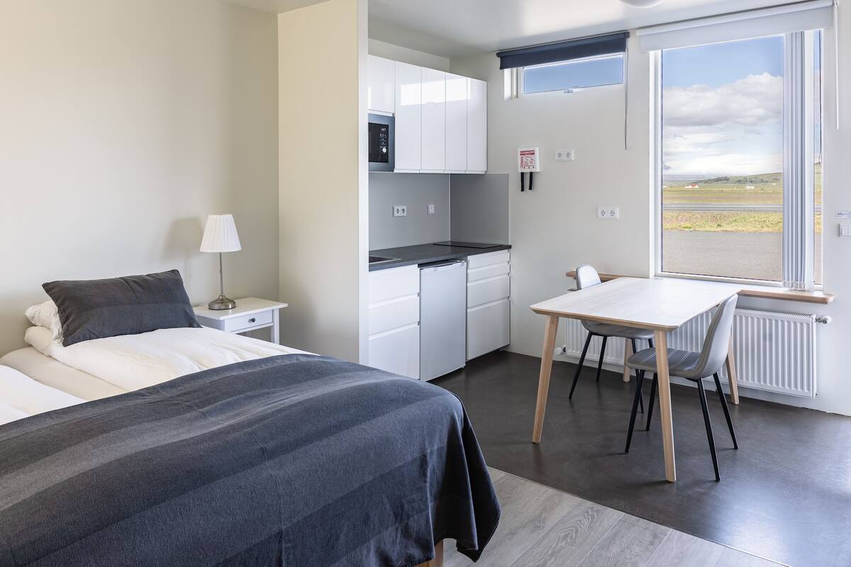 A bedroom with a bed, bedside table, and lamp. Adjacent is a kitchenette with a stove, sink, and cabinets. A dining table with chairs is near a window with a view of the outdoors.