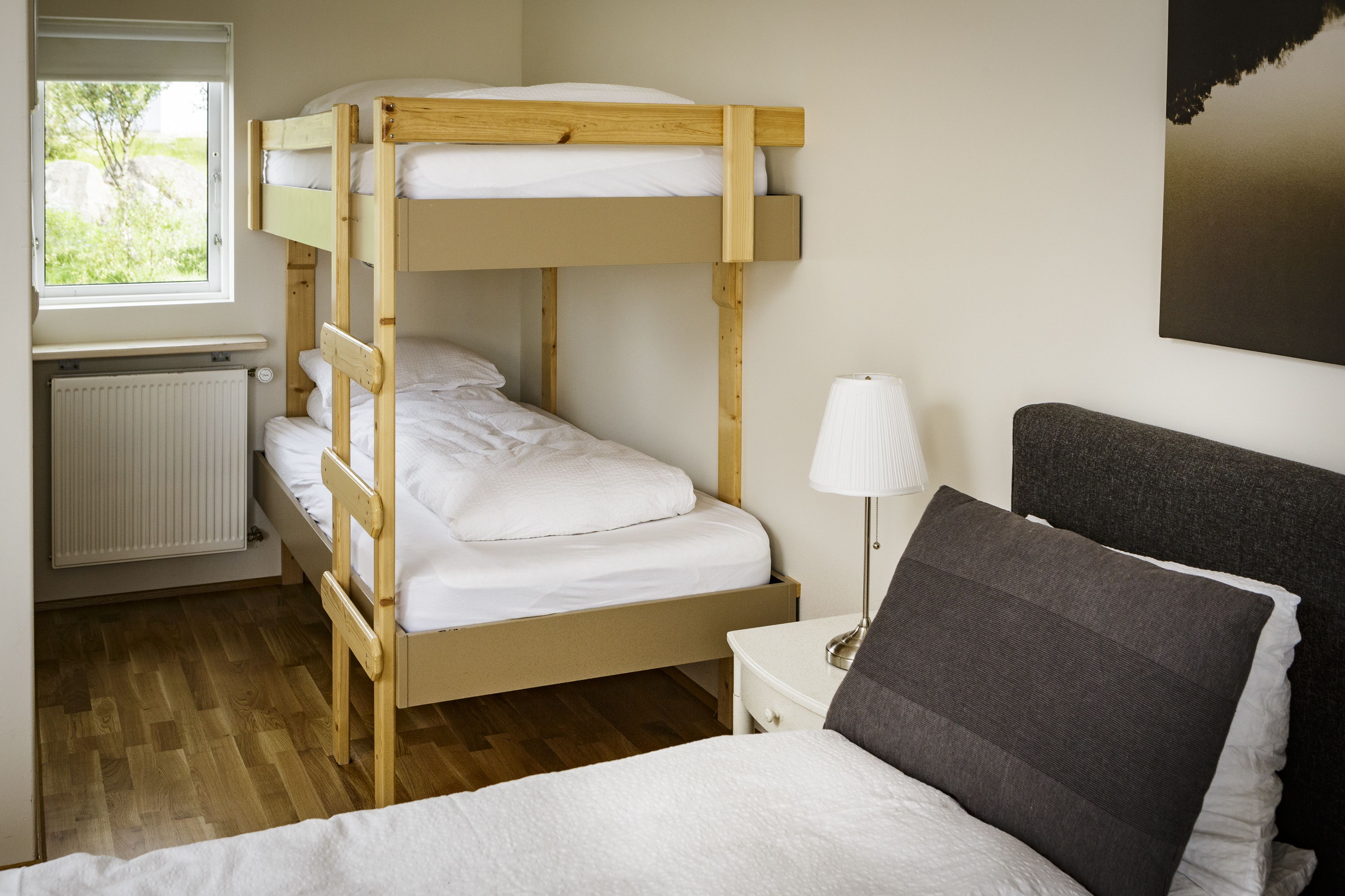 A bunk bed room with a window, radiator, bedside table, lamp, and a framed picture on the wall.
