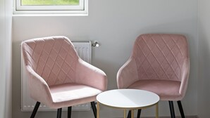 Two pink upholstered chairs with black legs and a small white table with gold legs, positioned in front of a window with a view of a tree and grass. - South Central Apartments (Selfoss)