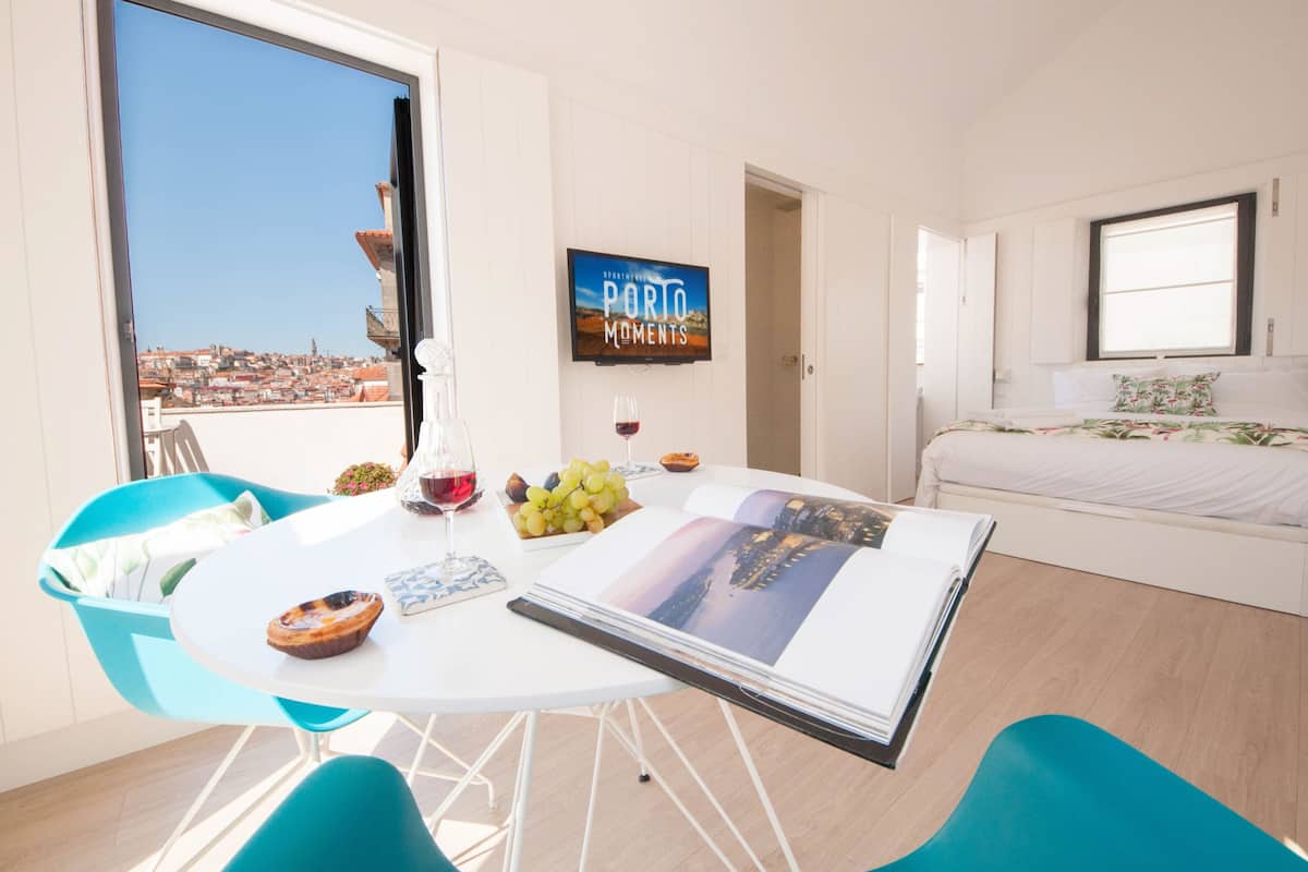 A modern living room with a large window, a dining area with a table set for two, and a bed with a floral bedspread.