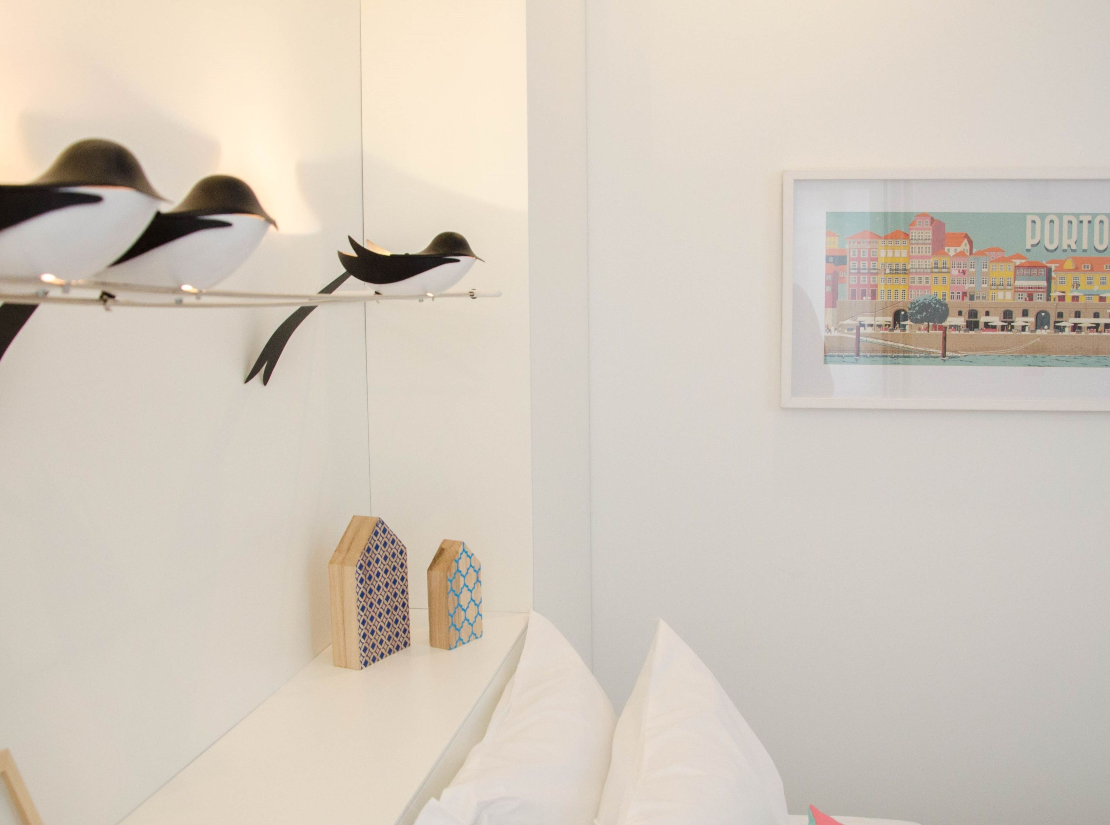 Un dormitorio con cama, almohadas, una estantería con objetos decorativos, una lámpara de pared y una fotografía enmarco de un paisaje urbano.