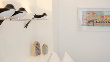 Un dormitorio con cama, almohadas, una estantería con objetos decorativos, una lámpara de pared y una fotografía enmarco de un paisaje urbano.