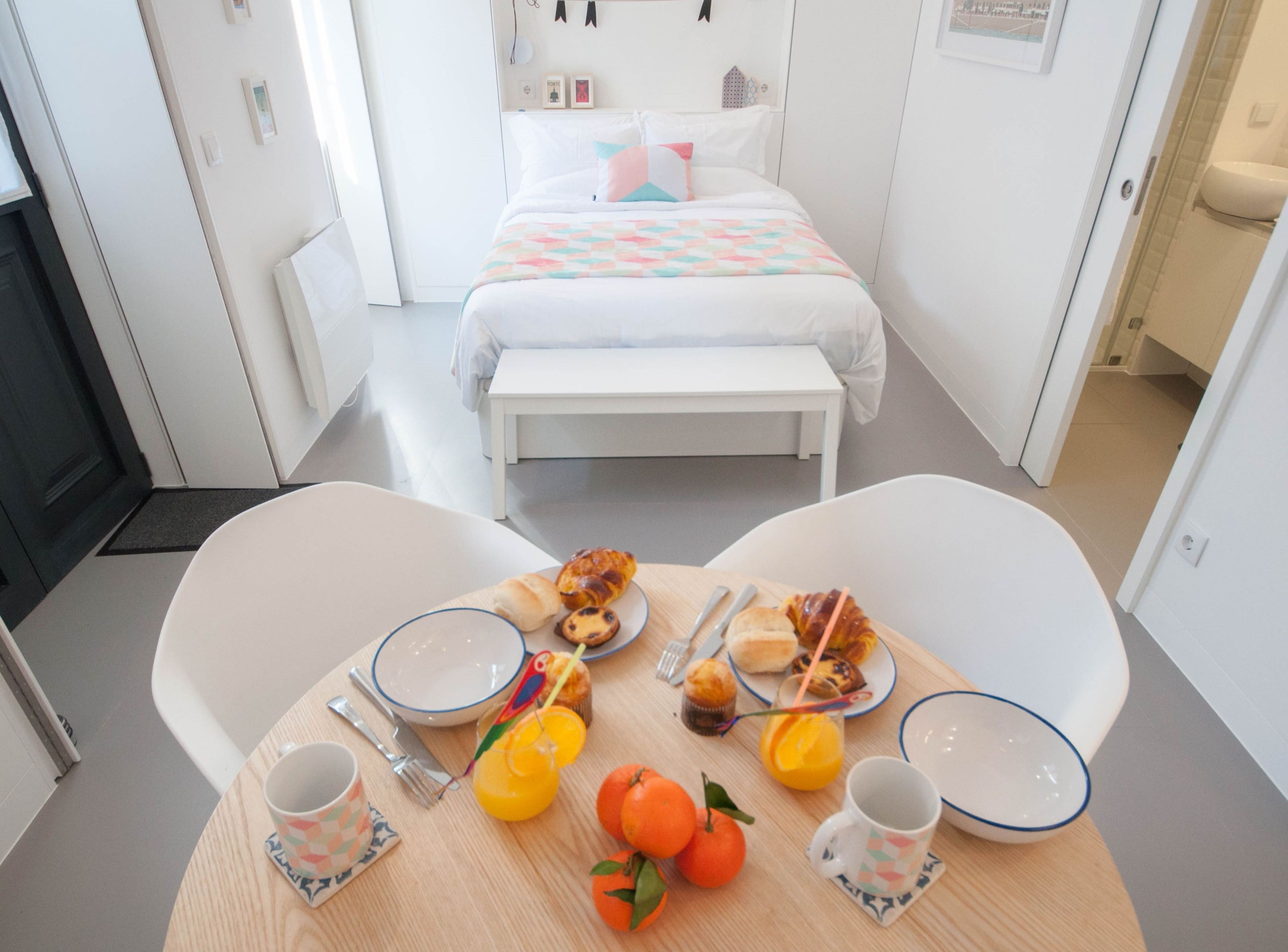 A hotel room with a neatly made bed, a dining table with breakfast items, and a bathroom visible in the background.