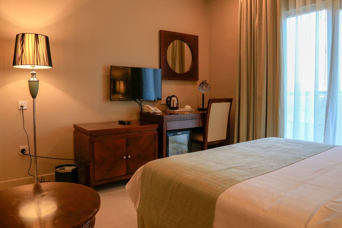 A hotel room with a bed, a desk with a computer, a television, a mirror, and a window with curtains.