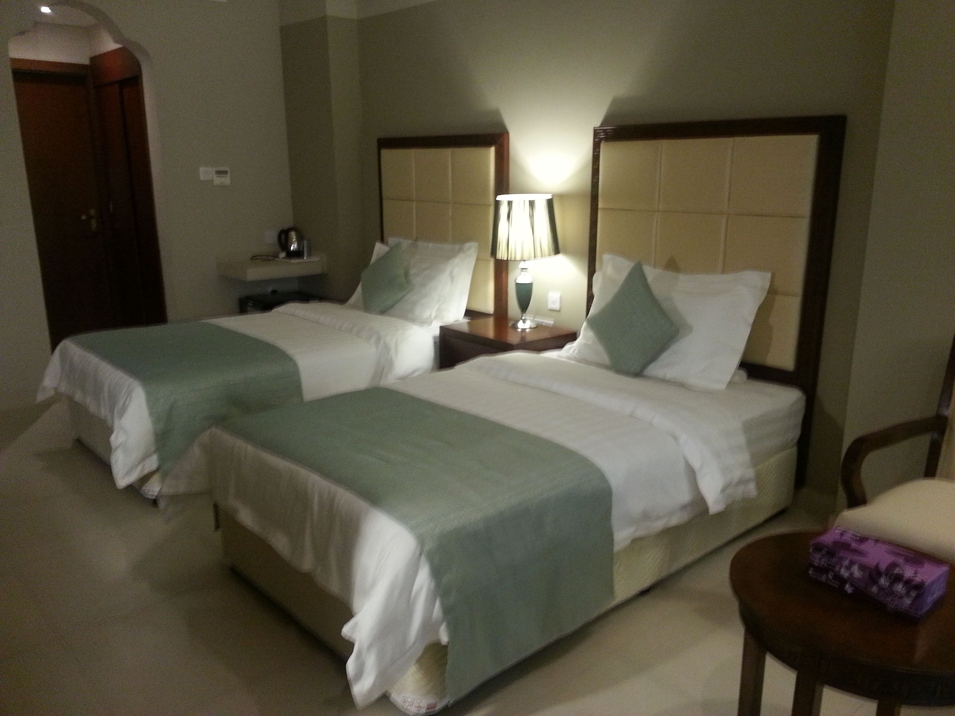 A hotel room with two single beds, a nightstand with a lamp, a chair, and a small table with a box.