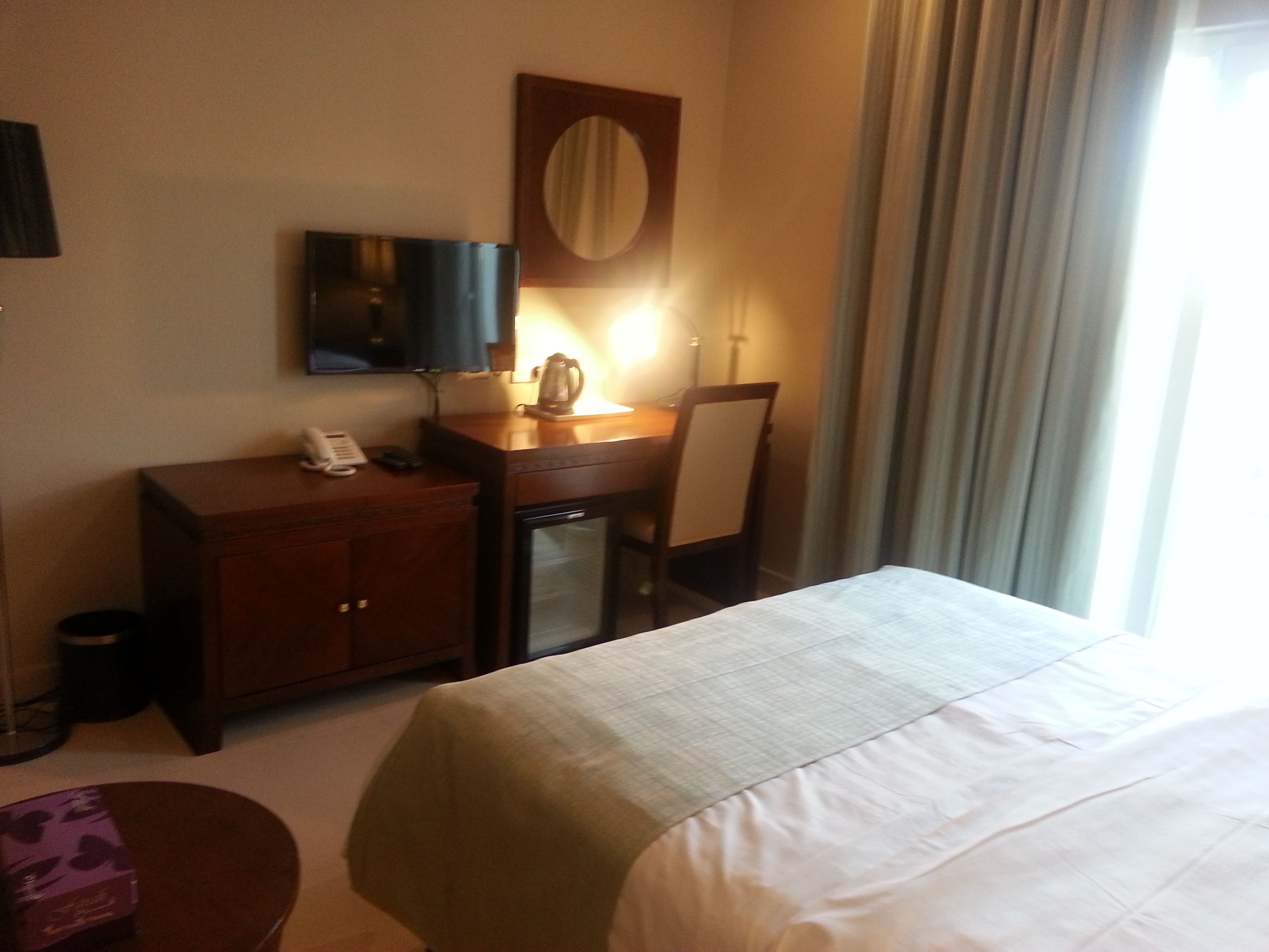 Habitación de hotel con cama, escritorio con teléfono, refrigerador pequeño, espejo y ventana con cortinas.