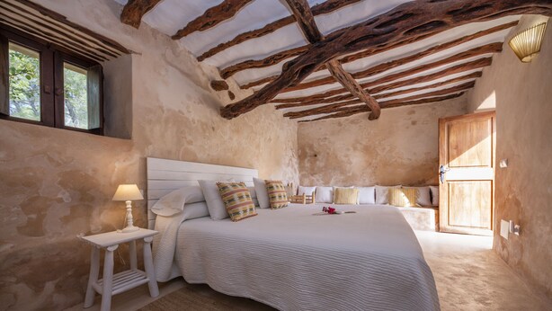 A bedroom with a large bed, wooden beams on the ceiling, a nightstand, and a window with blinds. - Agroturismo Can Cosmi Prats (Santa Eulalia del Rio)