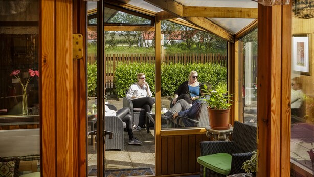 Een glazen conservatorium met houten kozijnen en deuren, met uitzicht op een tuin. Binnen staan rieten stoelen en een bijzettafeltje met een potplant.