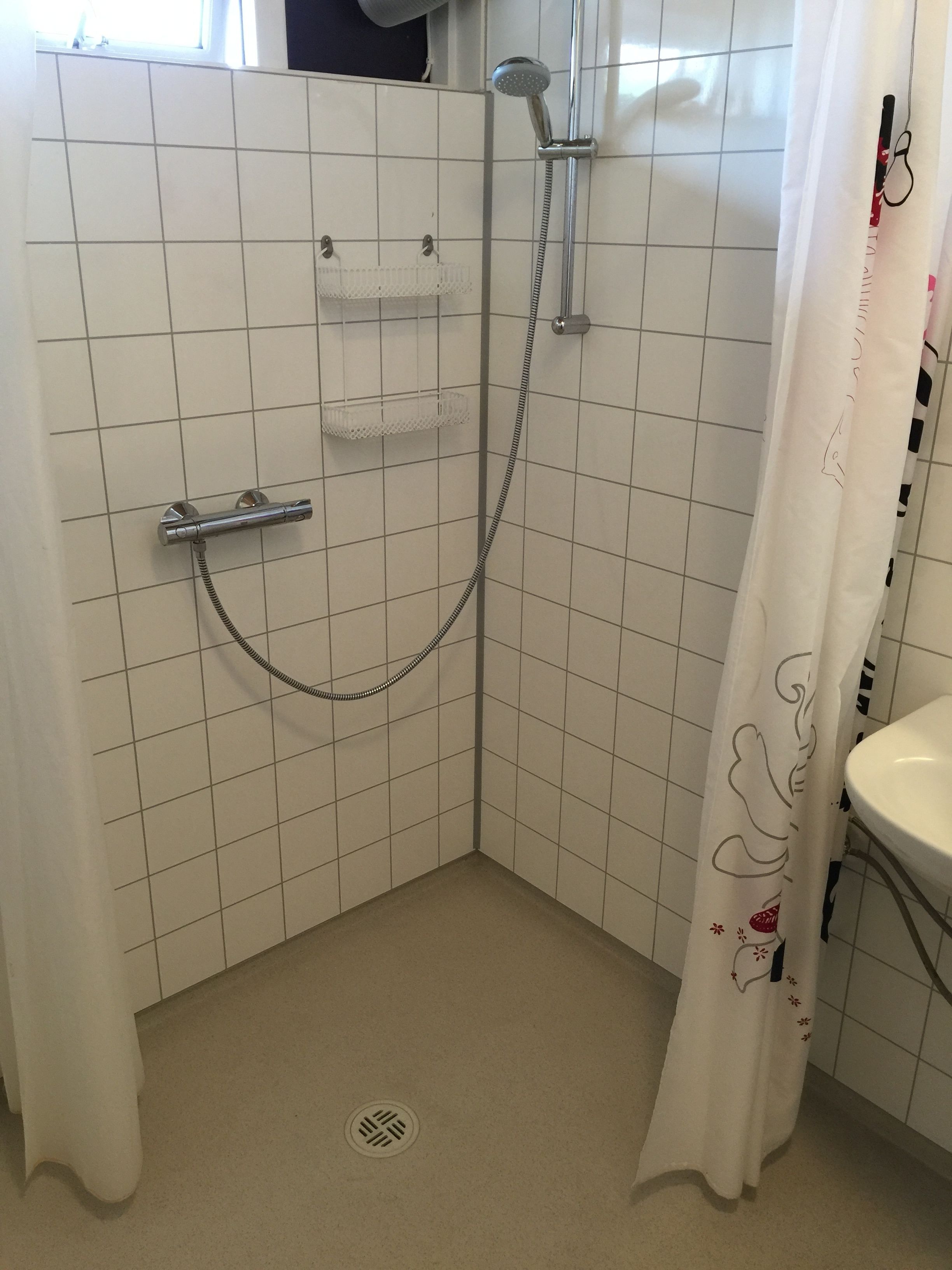 Un área de ducha pequeña, con azulejos, que incluye un cabezal de ducha manual y un grifo de pared. Se observa un estante pequeño encima del cabezal y un desagüe en el suelo.