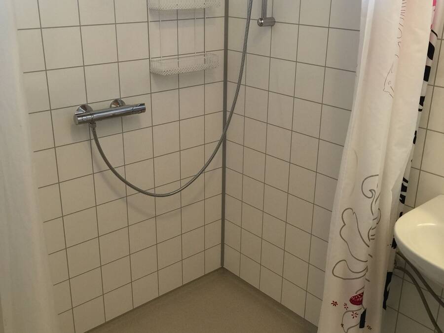 Un área de ducha pequeña, con azulejos, que incluye un cabezal de ducha manual y un grifo de pared. Se observa un estante pequeño encima del cabezal y un desagüe en el suelo.