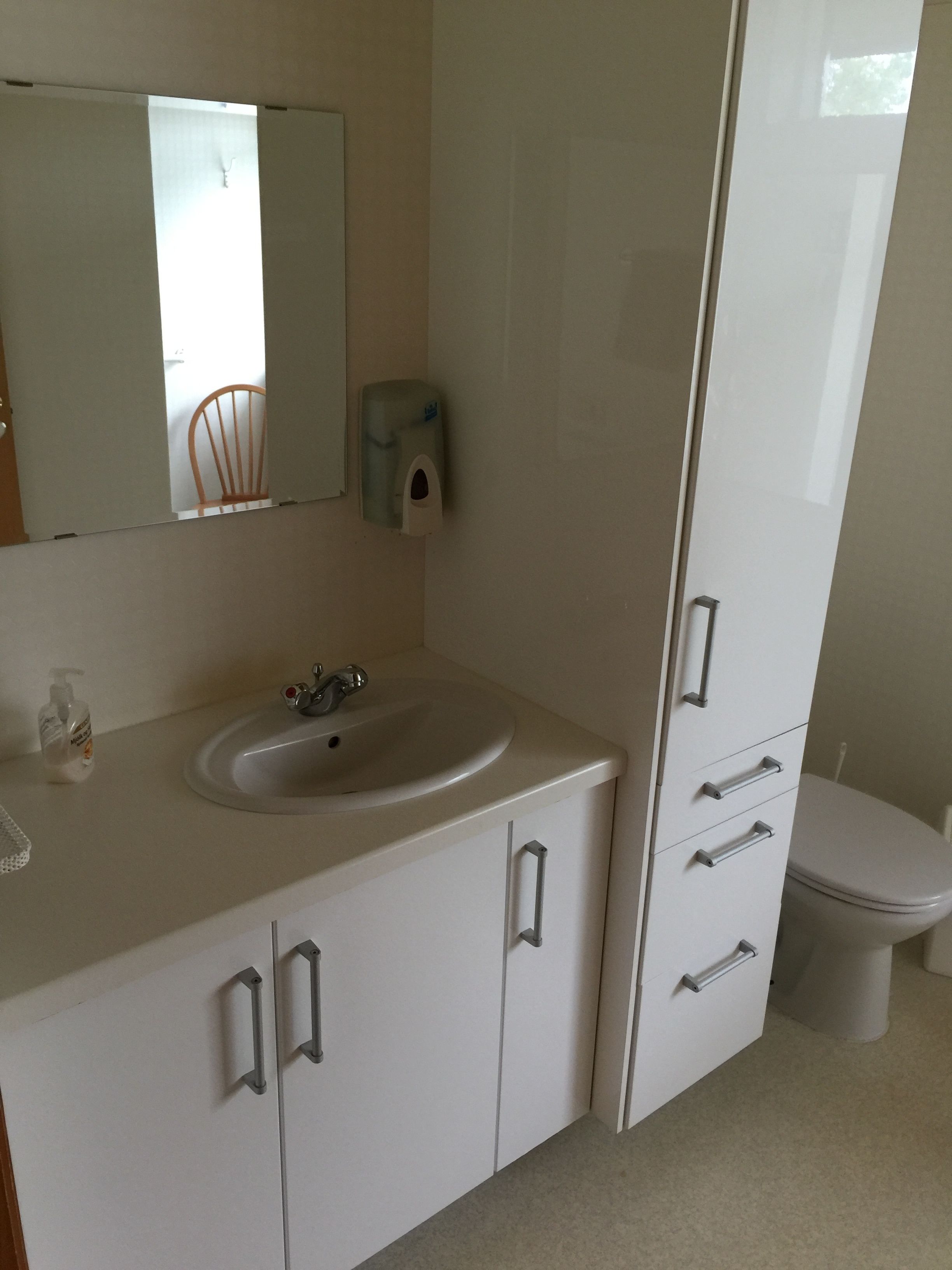 A bathroom with a white sink, mirror, and toilet. There is a soap dispenser on the wall and a bottle of hand soap on the counter.