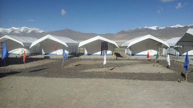 A row of white tents with blue and red flags, set against a mountainous backdrop. - Lotus Camp and Restaurant (Leh)