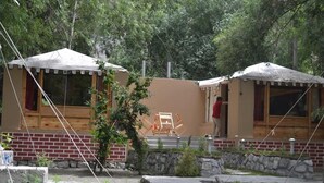 Duas cabanas de madeira com telhados de palha, uma parede de pedra e um banco de madeira.