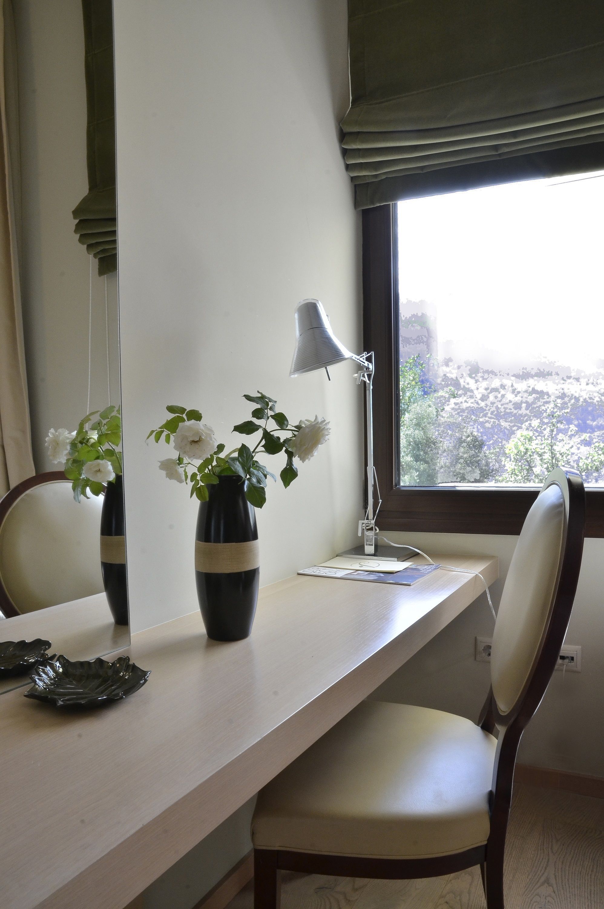 A desk with a lamp, a vase with flowers, and a chair in a room with a window.