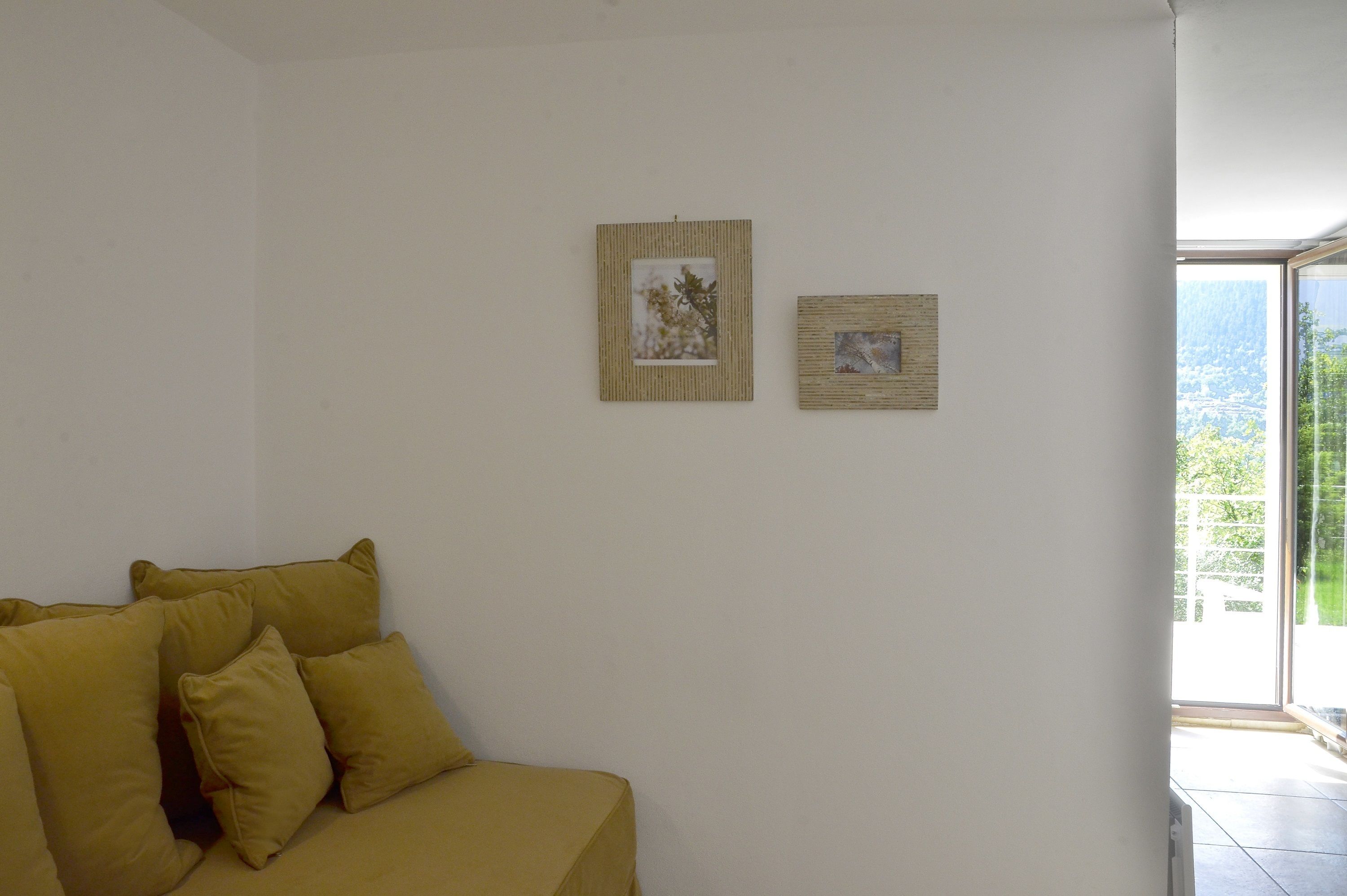 Una habitación con un sofá amarillo, dos cuadros en la pared y un ventanal con vistas a zonas verdes.