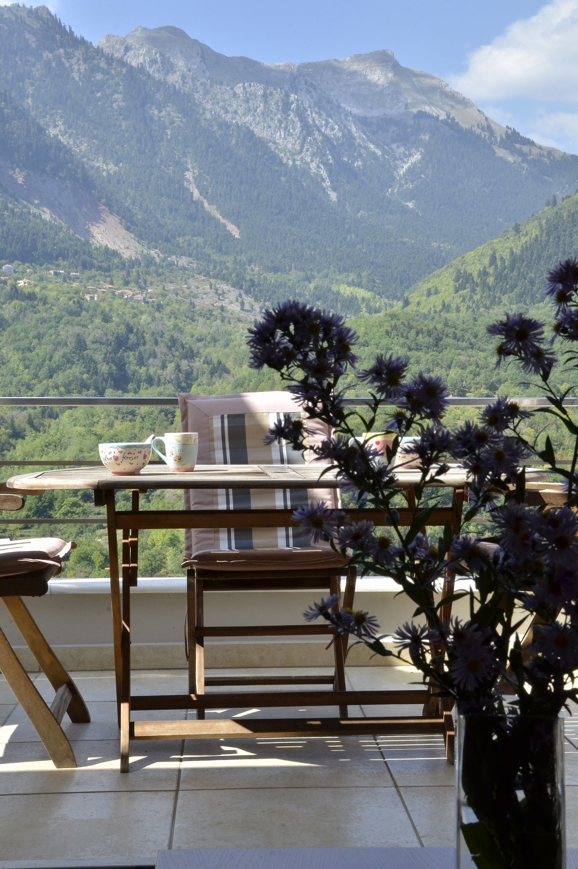 A balcony with a table, chairs, and a vase of flowers. A mountain view is visible in the background.