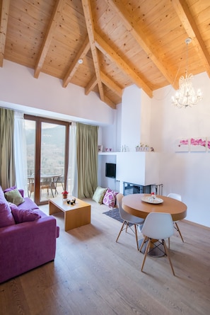 A spacious living room with a wooden ceiling, a round dining table with chairs, a purple sofa, and a view through a sliding glass door.