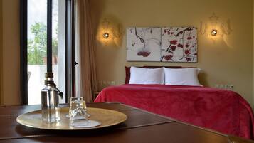 A hotel room with a bed, a bedside table with a bottle and glasses, a wall-mounted painting, and two wall sconces.