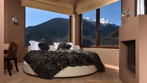 Een slaapkamer met een rond bed, een stoel, een open haard en een groot raam met uitzicht op de bergen.