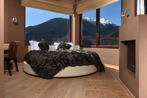 A bedroom with a round bed, a chair, a fireplace, and a large window with a mountain view.