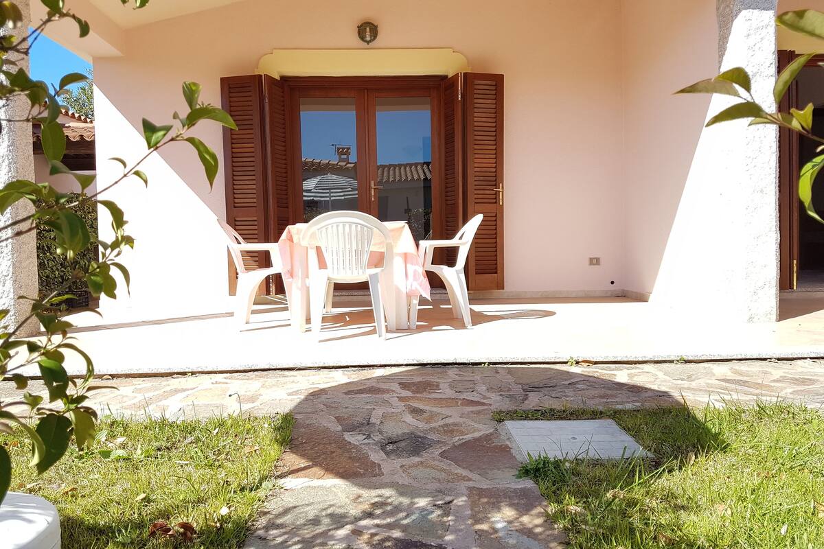 Uma área de estar coberta ao ar livre, com mesa e cadeiras, uma porta de madeira e um caminho de pedra que leva à entrada.
