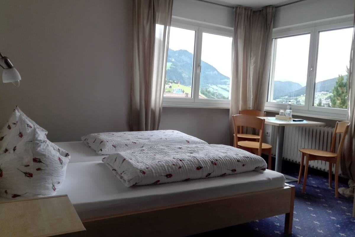 Une chambre d’hôtel avec un lit, un bureau et une chaise. Il y a une fenêtre donnant sur les montagnes.