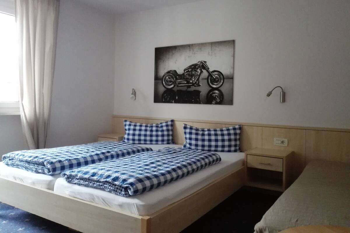 Un lit double avec une literie à carreaux, une table de chevet, une commode, une œuvre d’art représentant une moto accrochée au mur, et une fenêtre avec des rideaux.