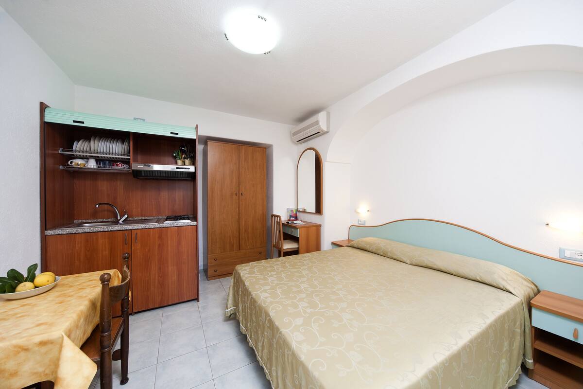 Ein Schlafzimmer mit einem Bett, einem Nachttisch, einem Schreibtisch mit Stuhl, einem Kleiderschrank und einer Küchenzeile mit Spüle und Regalen.