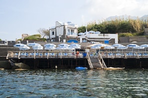 A pier with blue umbrellas and lounge chairs, a staircase leading down to the water, and a building with a balcony in the background. - La Rotonda Sul Mare (Forio)